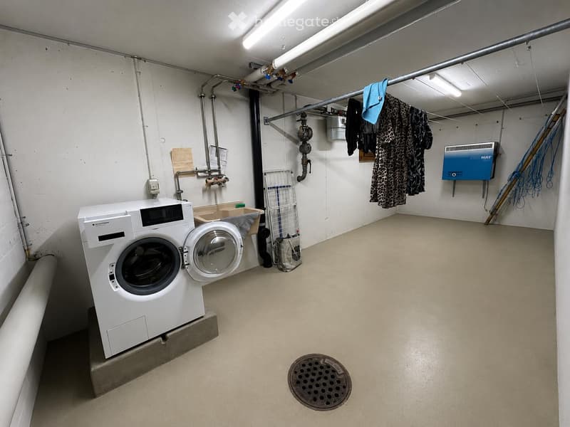 Laundry room with a washing machine, dryer, laundry basket, and laundry rack. The room has exposed pipes and a drain in the floor.