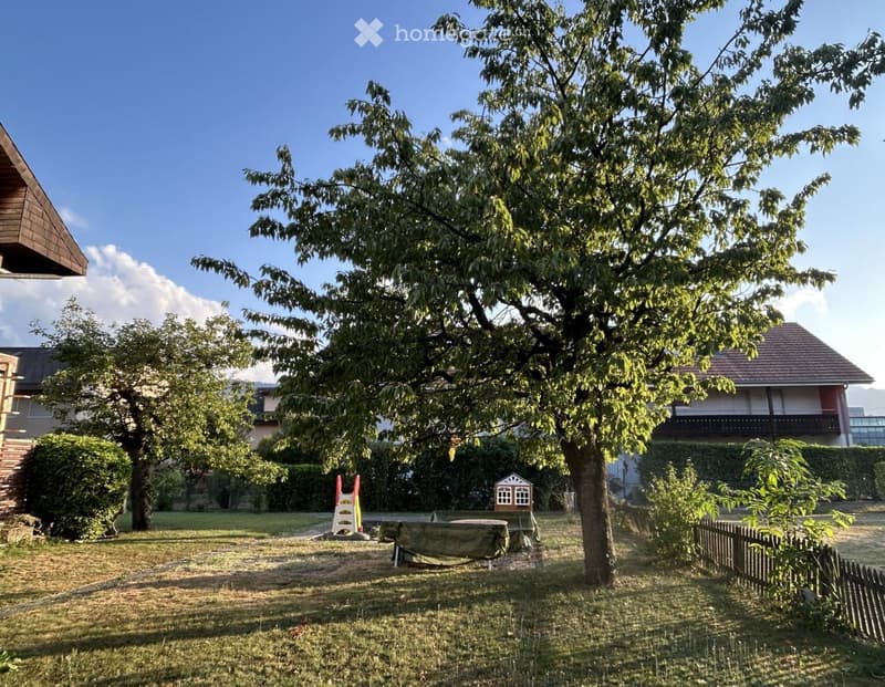 A well-maintained garden with a large tree, a covered table, a small playhouse, and a wooden fence. The area is surrounded by houses and greenery.