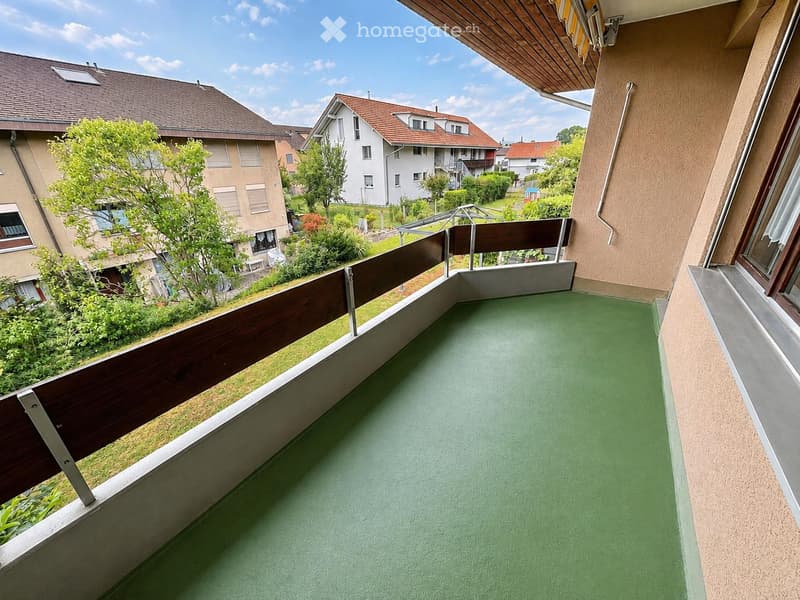Balcony with green flooring, wooden railing, overlooking a residential area with houses and greenery.