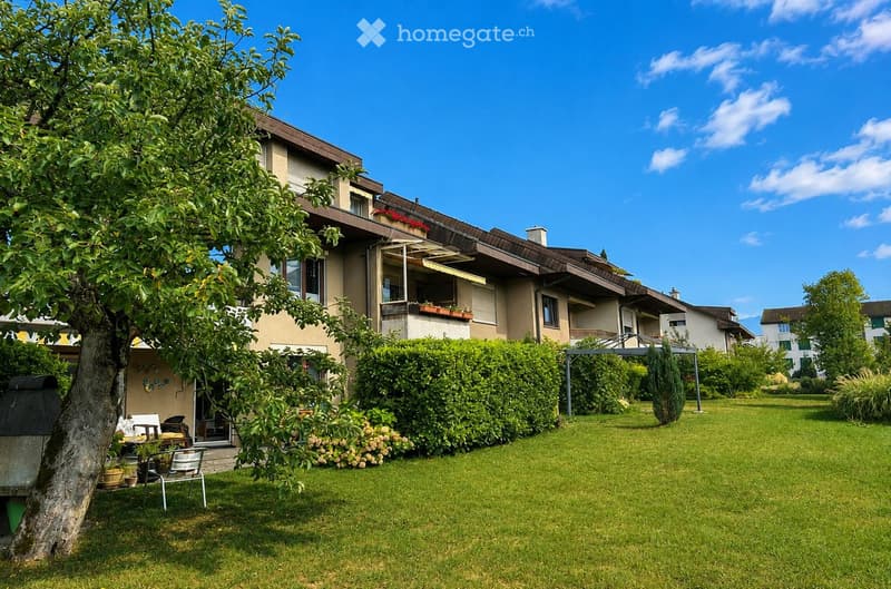 Well-maintained garden with a lawn, trimmed hedges, patio furniture, and a tree, adjacent to a multi-story residential building with balconies and windows.