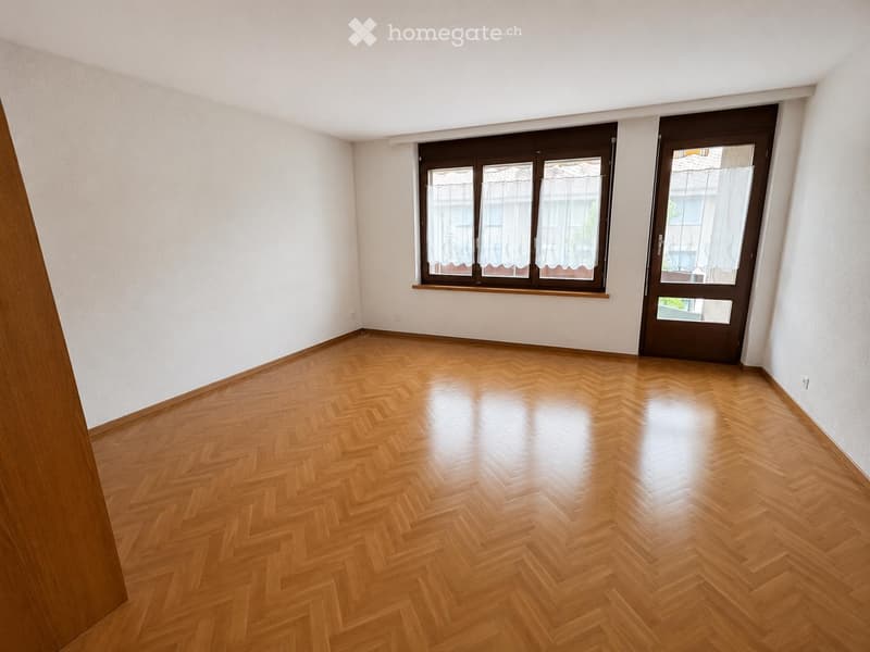 Empty room with wooden herringbone flooring, white walls, large window with brown frame, and a dark brown door.
