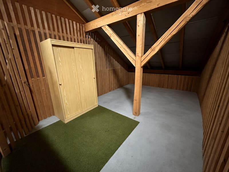 An unfurnished attic space with wooden walls, exposed wooden beams, and a mix of carpeted and concrete flooring.