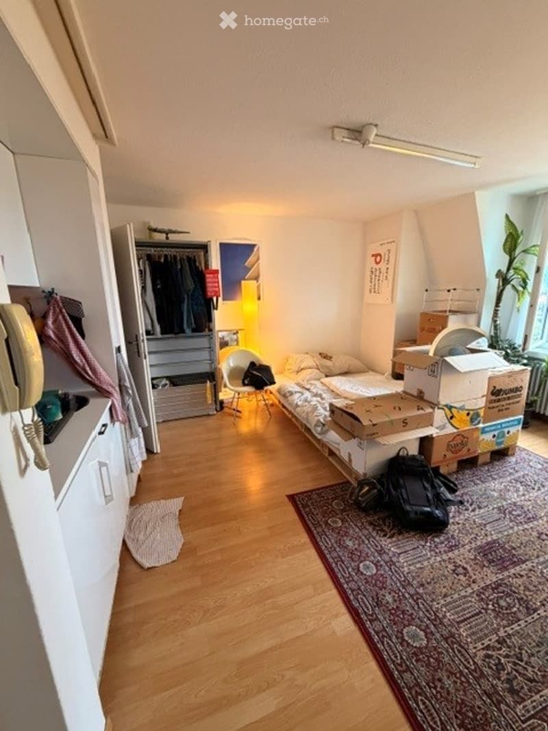A small, cozy room with a bed, a chair, a wardrobe, and a rug. The room has wooden flooring and is partially packed with boxes, indicating it might be in transition.