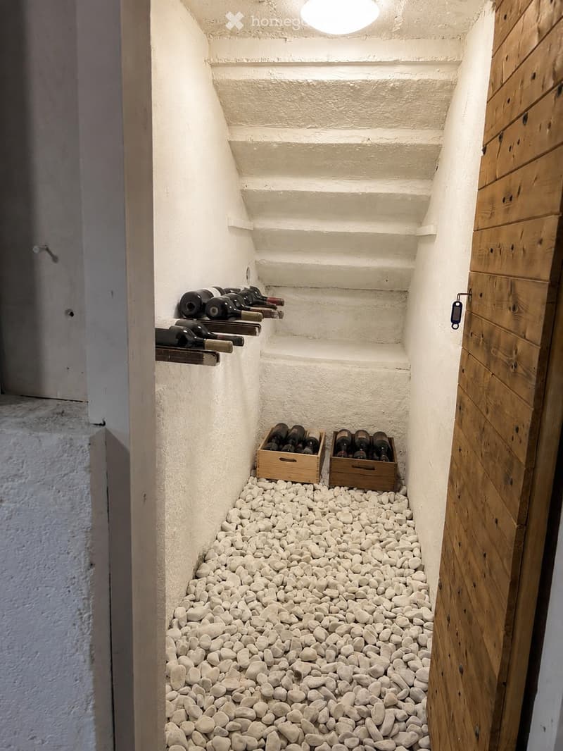 Narrow hallway with white textured walls, wooden door, and white pebble flooring. Wine bottles are stored on shelves and in wooden crates along the walls.