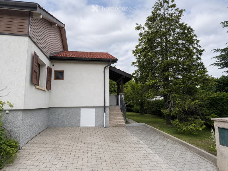 Two-story house with a tiled roof, white exterior, and a covered entrance with steps leading up to it. The house has brown shutters and a small window on the upper level. The surrounding area includes a paved driveway and greenery.