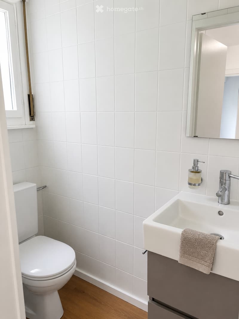 Modern bathroom with white tiled walls, a white toilet, a white sink with a modern faucet, a mirror above the sink, and a towel on the sink counter.
