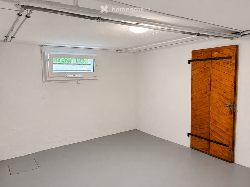 Empty room with white walls, grey floor, wooden door, and small window showing greenery outside.