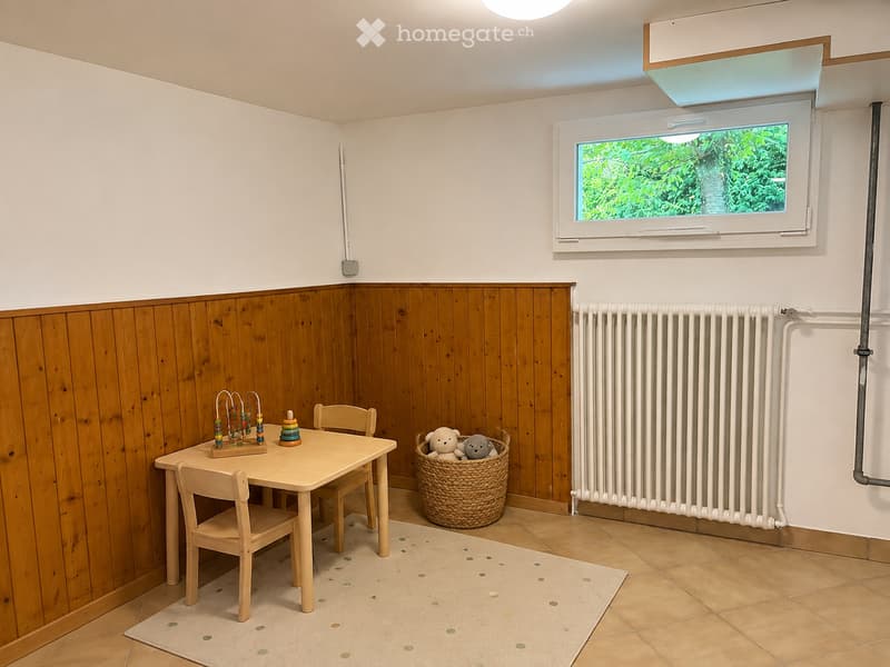 A small, cozy room with wooden paneling on one wall, a table with chairs, a basket with stuffed toys, and a radiator.