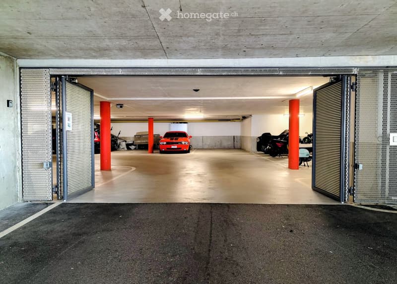 Underground parking area with several parked vehicles, including a red sports car, motorcycles, and other cars. The space features concrete pillars, a clean and organized layout, and metal security gates at the entrance.