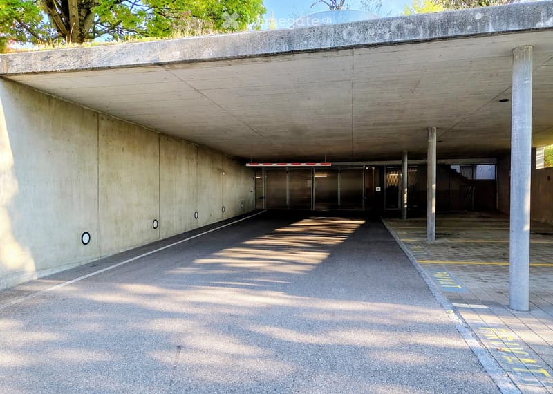A modern underground parking area with concrete walls and a covered entrance. The area features a smooth concrete floor, several support pillars, and a gated entrance leading to the upper level.