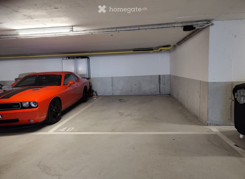 Underground parking space with a bright orange sports car parked in bay number 40. The area features concrete floors and walls, fluorescent lighting, and a storage cabinet in the background.