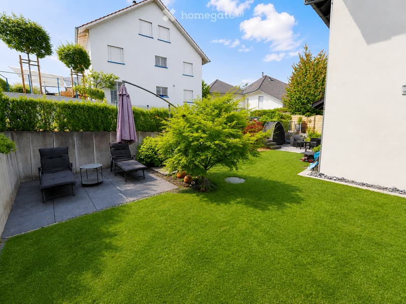 Well-maintained garden with a neatly trimmed lawn, a small patio area with lounge chairs and a table, and surrounding greenery including bushes and trees. A modern house is visible in the background.