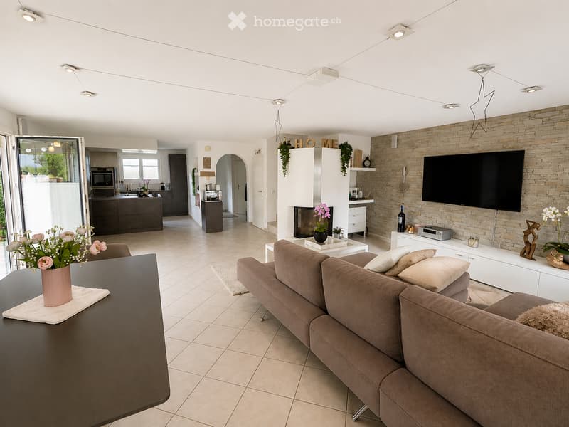 Modern living room with a brown sofa, coffee table, fireplace, and television. The space features a kitchen area with a microwave and cabinets, and decorative elements like vases and flowers.