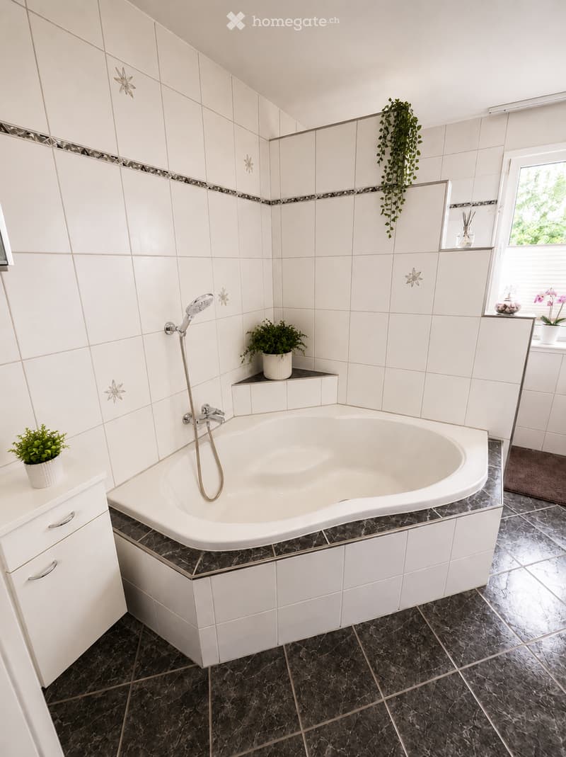 Modern bathroom with a large bathtub, white tiles, dark marble flooring, and decorative potted plants on the cabinet.