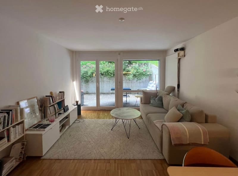 Bright living room with wooden flooring, large sliding glass doors leading to a balcony, a beige sofa with multiple cushions, a round coffee table, a bookshelf, and a small table with a chair.