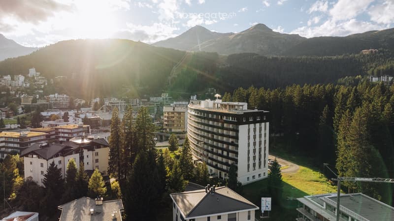 Aerial view of a town nestled in a mountainous landscape, featuring a mix of residential and commercial buildings surrounded by dense forests and snow-capped peaks.