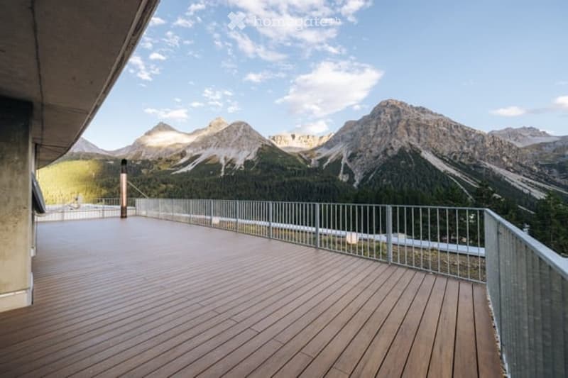 Wooden deck with metal railing, overlooking a scenic mountain range with snow-capped peaks and green valleys.