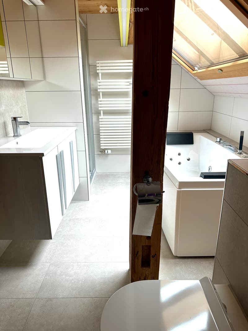 Modern bathroom with a sink, bathtub, radiator, mirror, and cabinets. Features a skylight window and wooden beam.