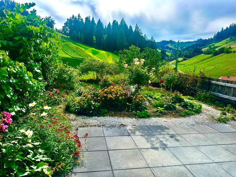 A well-maintained garden with a variety of colorful flowers, shrubs, and trees, set on a paved patio with a scenic view of rolling green hills and a partly cloudy sky.