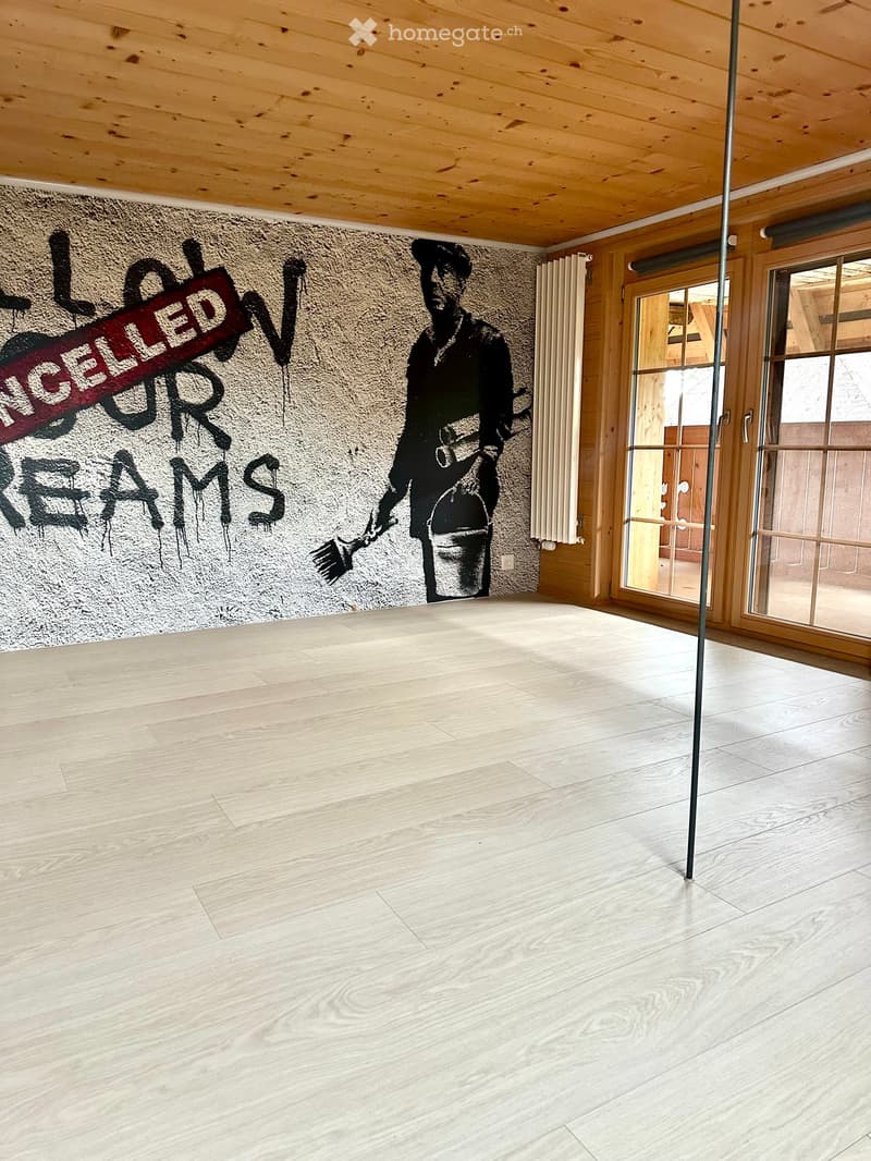 Empty room with light wooden flooring, exposed wooden ceiling beams, and a large mural on one wall. The room features large windows and a door leading outside.