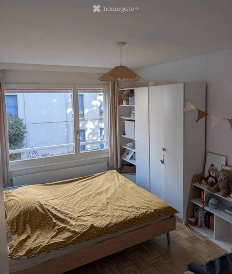 Bedroom with a wooden bed covered in a yellow duvet, a white wardrobe, a bookshelf with books and a teddy bear, and a hanging lamp above the bed.