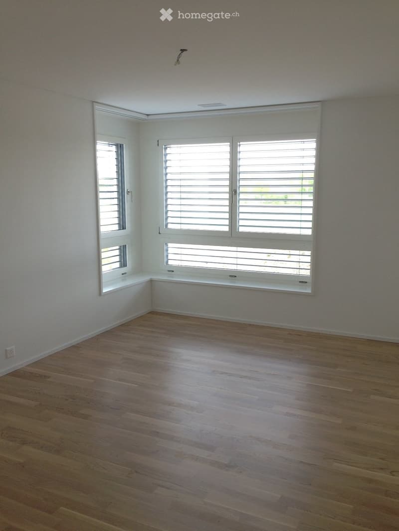 Empty room with wooden flooring, white walls, and large windows with blinds.