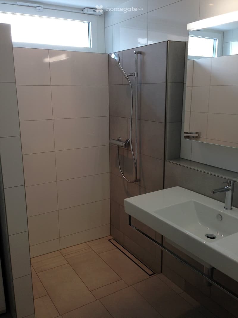 Modern bathroom with beige tiled walls, a white sink with a chrome faucet, a showerhead, and a large mirror above the sink.