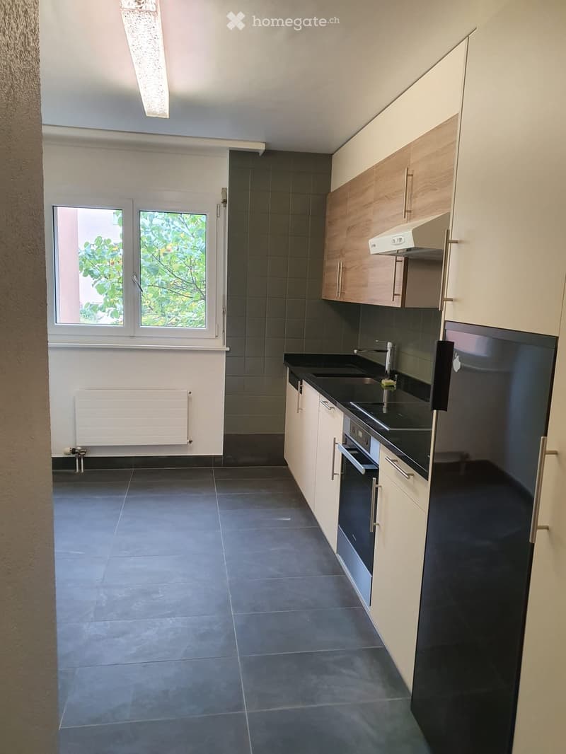Modern kitchen with white and wood cabinets, black countertops, stainless steel appliances, and large windows with a view of greenery.