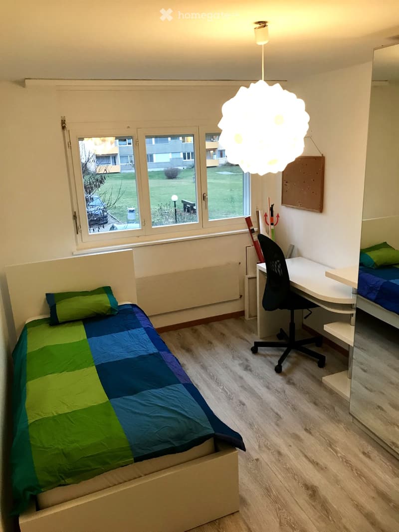 Cozy bedroom with a single bed, desk, chair, and a hanging lamp. The bed has a green and blue quilt, and the room features wooden flooring and large windows with a view of the outdoors.