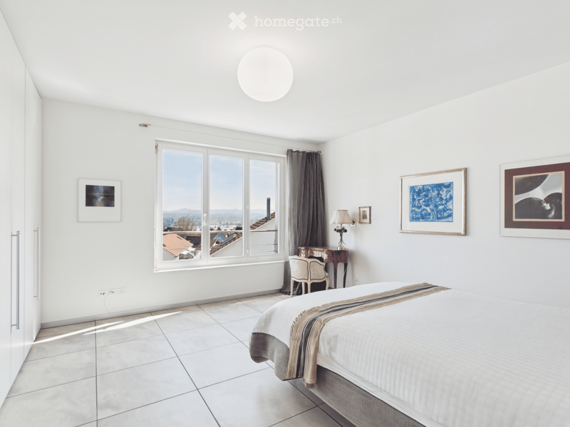 Bright, modern bedroom with white walls, large windows offering a city view, a neatly made bed, a small table with a lamp, and several picture frames on the walls.