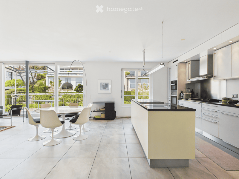 Modern kitchen with white cabinets, a central island, and a dining area with white chairs and a round table. Large windows provide natural light, and the space features contemporary appliances and sleek design elements.