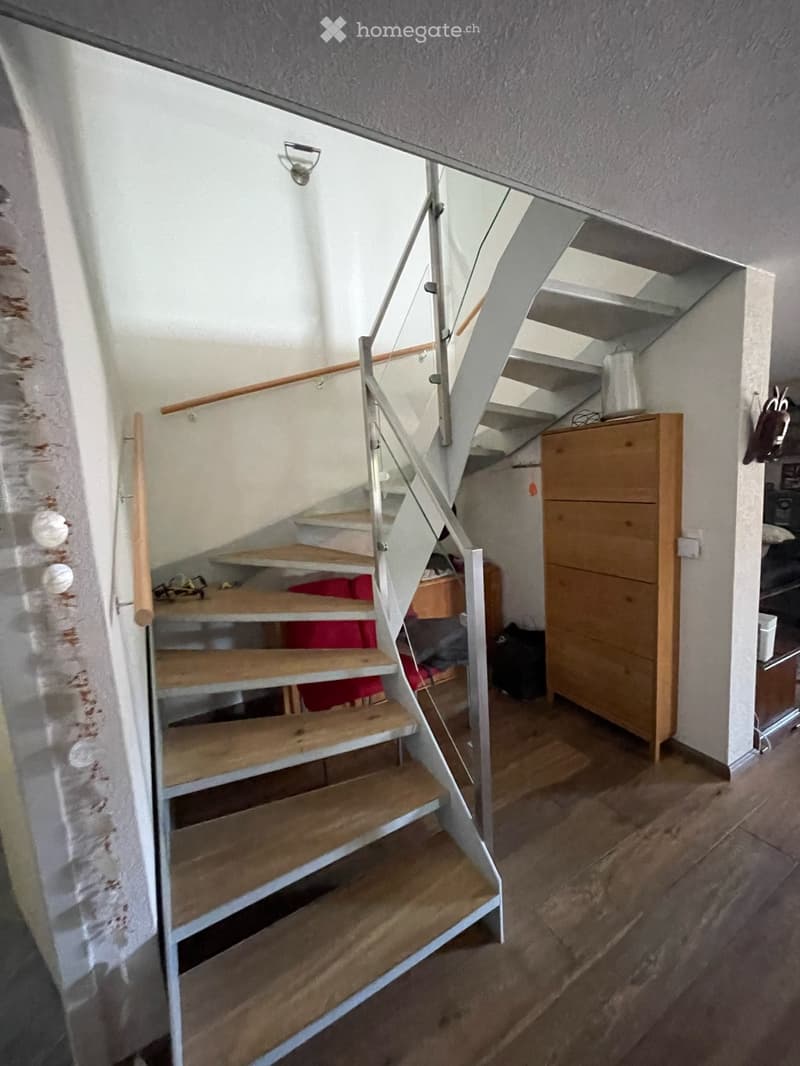 Wooden staircase with metal railing, wooden floor, white walls, and a small wooden cabinet in the background.