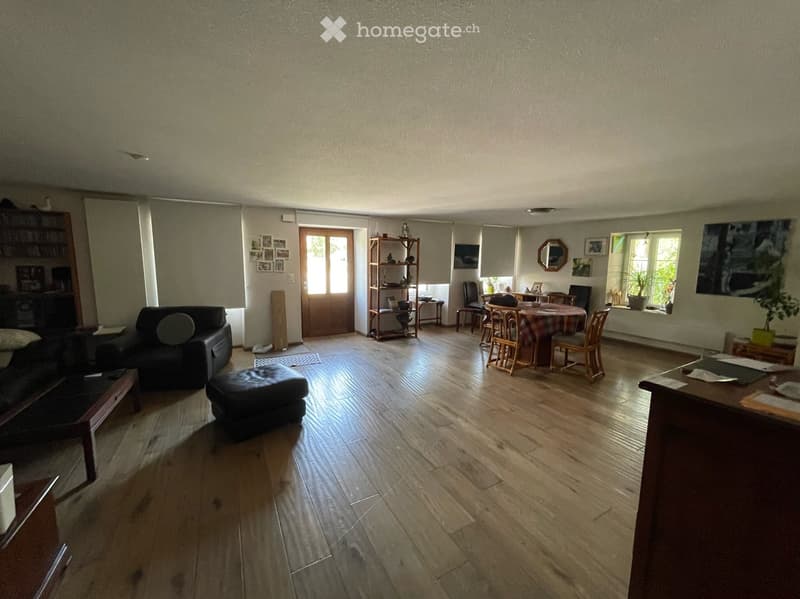 Open-plan living and dining area with wooden flooring, a black leather sofa, ottoman, coffee table, dining table with chairs, shelves, and a mirror on the wall.