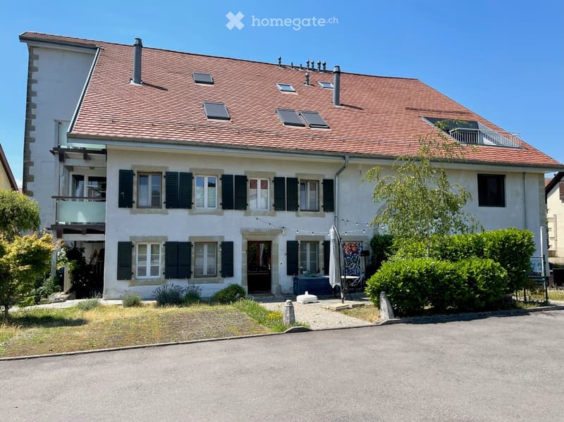 Two-story house with red-tiled roof, white exterior, multiple windows with shutters, balcony on the upper level, and a small garden area with patio furniture.
