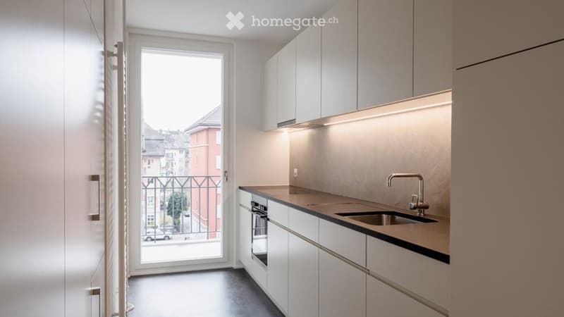 Modern kitchen with white cabinets, stainless steel sink, and built-in oven. Features a large window with a view of the street and a balcony.