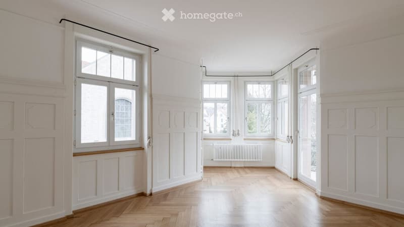 Completely empty room with white walls, wooden floor, large windows, and wainscoting.