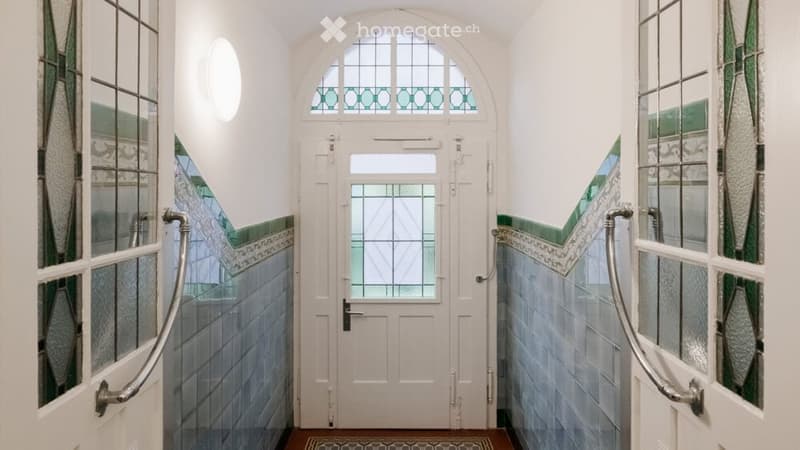 A narrow hallway with a white door at the end, featuring decorative stained glass windows and a patterned tile floor.