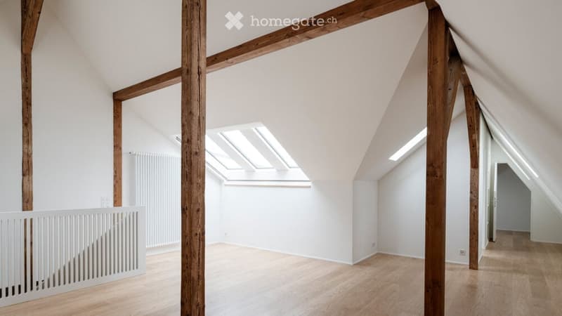 Bright, open attic space with wooden beams, skylights, and wooden flooring. The room is completely empty, with white walls and a white staircase railing visible.