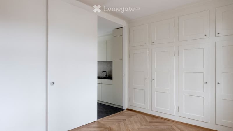 Empty room with wooden flooring, white built-in wardrobes, and an open doorway leading to a small kitchen area.