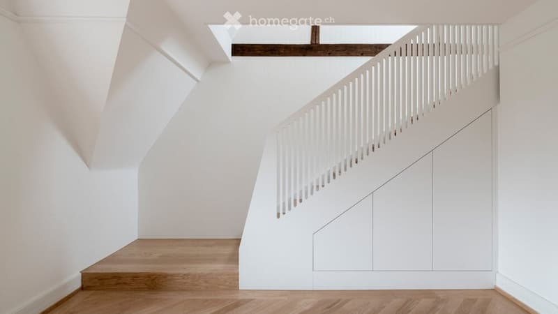 A modern, minimalist hallway with white walls, wooden flooring, and a white staircase with a wooden handrail. The space features built-in white cabinetry along one wall and a skylight above.