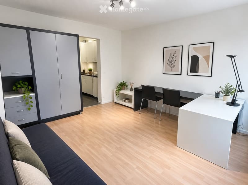 Modern, well-lit room with a sofa, desk, and cabinet. Features a kitchen area in the background with white cabinets and a countertop.