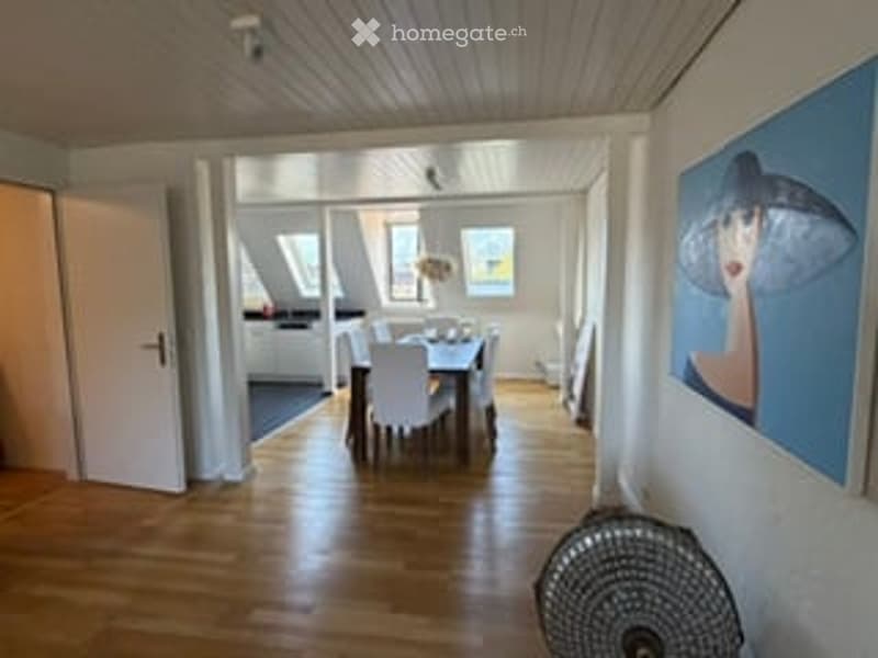 Bright dining area with wooden flooring, white walls, and large windows. A dining table with chairs is centered, and a fan is visible on the floor.