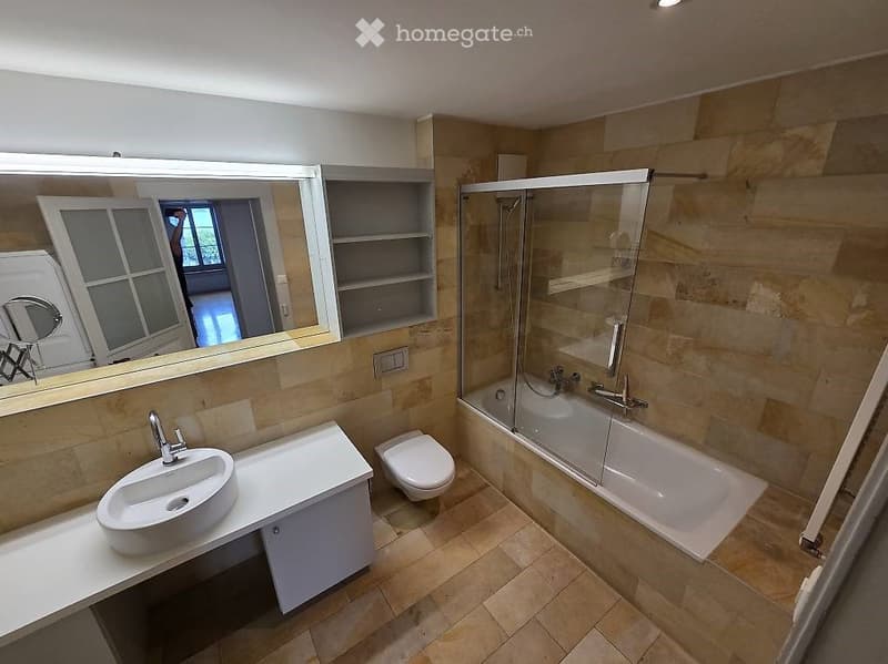 Modern bathroom with beige tiles, a large mirror, a sink with a modern faucet, a toilet, a bathtub with a glass shower enclosure, and built-in shelves.