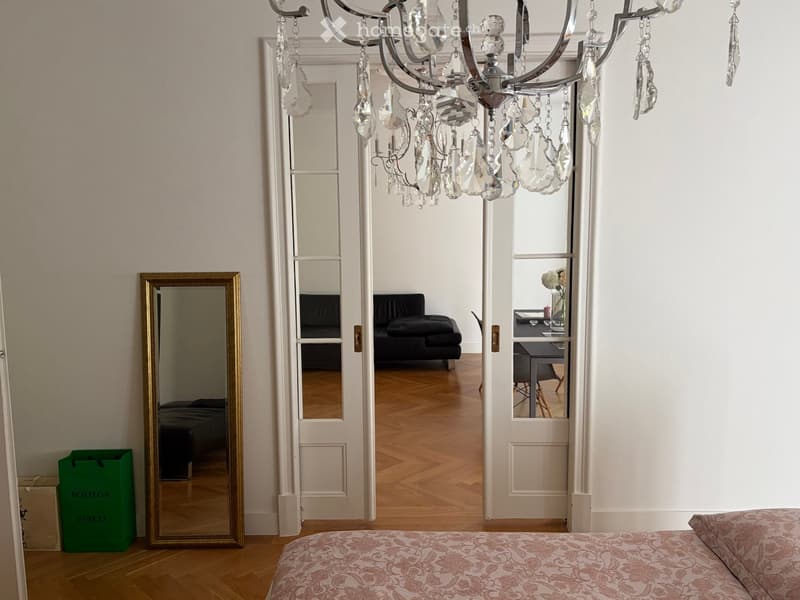 Elegant hallway leading to a furnished room with a bed, sofa, chair, table, and a large mirror. A chandelier hangs from the ceiling.