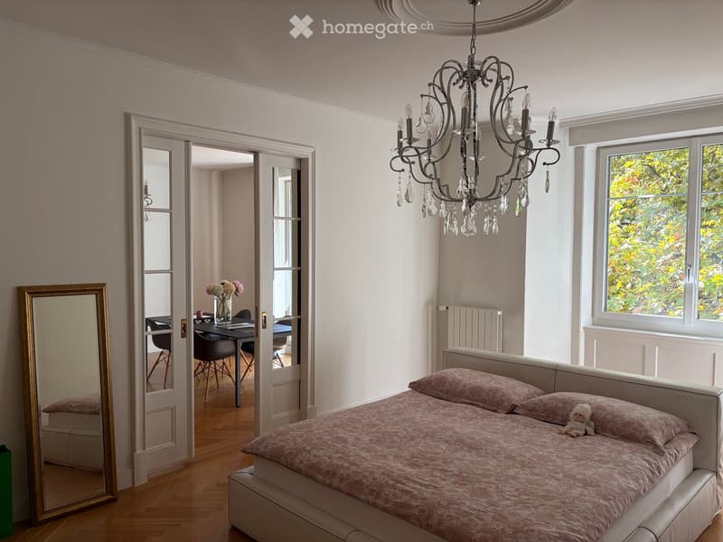 Elegant bedroom with a large bed, chandelier, and a mirror. A small table and chairs are visible in the adjacent room.