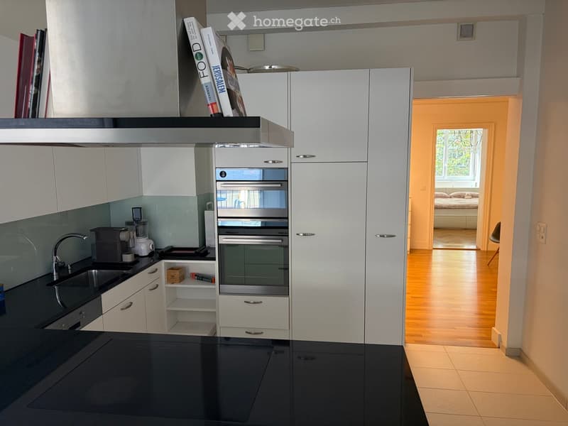 Modern kitchen with white cabinets, black countertops, stainless steel appliances, and a visible bedroom area in the background.