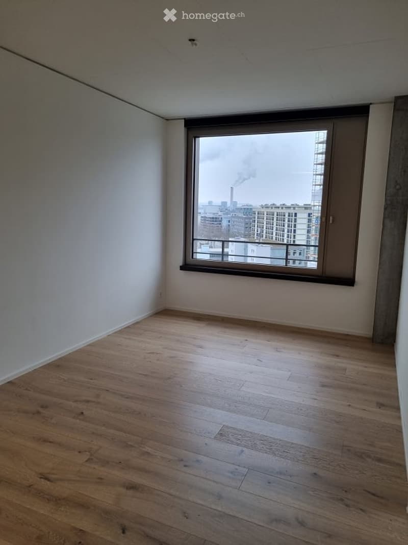 Empty room with wooden flooring and large window offering city view.
