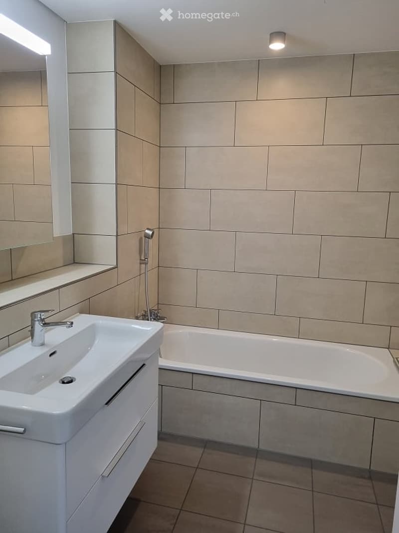 Modern bathroom with a white sink, bathtub, and large mirror. Beige tiles on walls and floor, sleek fixtures, and bright lighting.