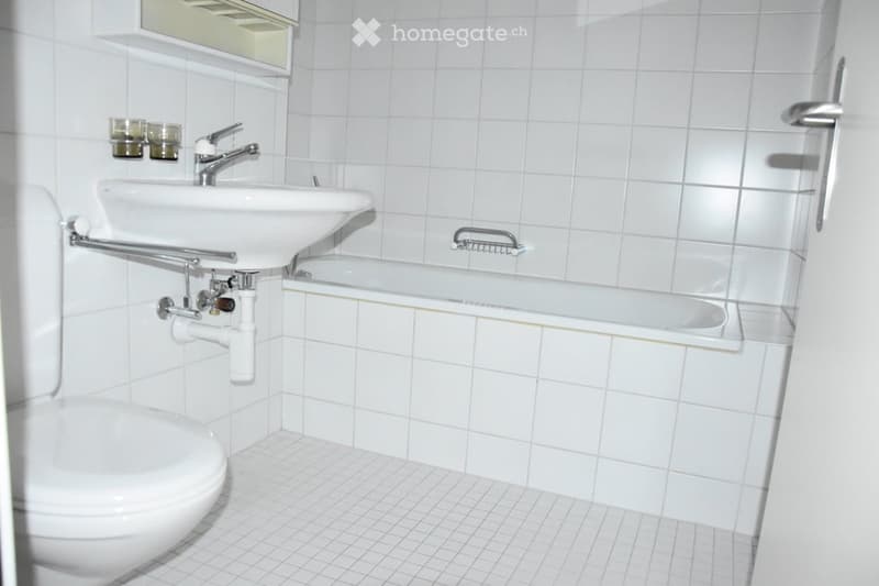 A small bathroom with white tiled walls and floor, featuring a toilet, sink with a faucet, a bathtub, a mirror, and a handrail for support.
