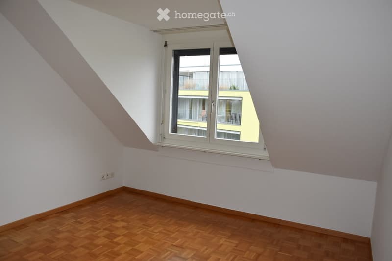 Empty attic room with wooden flooring, white walls, and a large window offering a view of neighboring buildings.